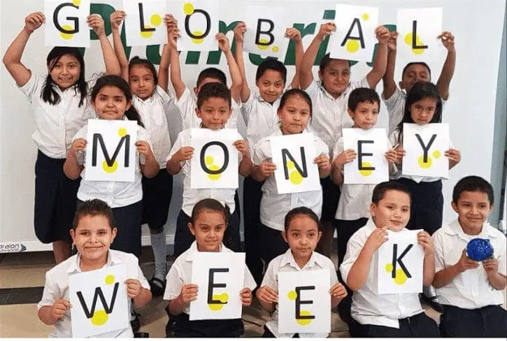 A group of schoolchildren in uniform are holding up paper signs with letters that spell GLOBAL MONEY WEEK, celebrating Global Money Week 2025. They are standing together and smiling for the photo.